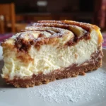 A close-up slice of creamy cinnamon roll honeybun cheesecake with a golden graham cracker crust, thick velvety cheesecake layer, and a beautiful cinnamon sugar swirl on top, lightly dusted with powdered sugar.