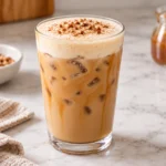 Iced pecan sticky bun latte in a clear glass with caramel drizzle, brown sugar cold foam, chopped pecans, and cinnamon.